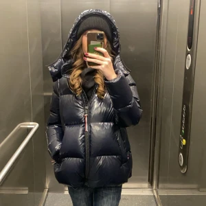 Mörkblå pufferjacka med huva - Säljer en snygg mörkblå tommy hiltfiger  pufferjacka med glansig yta och stor huva. Jackan har dragkedja framtill och breda, quiltade sektioner som ger en cool och modern look. Perfekt för kalla dagar och enkel att styla med mössa och jeans. Köpt för ca 2000