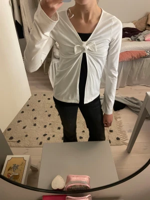 Vit tröja med rosett från H&M - Supersöt vit tröja från H&M med lång ärm och en  rosett framtill. Koftan har v-ringad hals och är gjord i mjuk bomull, perfekt att slänga över en topp. Enkel och clean stil som funkar till många outfits.