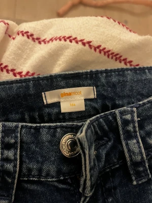 Blå bootcut jeans från Gina Tricot - Snygga blå jeans från Gina Tricot i storlek 164. Modellen har bootcut-ben och klassisk femficksdesign. Baksidan har coola broderade detaljer på fickorna med knappar. Jeansen är tillverkade i jeansmaterial och har normal passform.