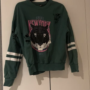 Grön sweatshirt med tigertryck Gina Tricot - Cool grön sweatshirt från Gina Tricot med stort tigertryck och texten 'Bronx New York' i rosa och vitt framtill. Tröjan har vita ränder på ärmarna och slitna detaljer med hål. Mjuk bomullskänsla och långärmad modell, perfekt för en streetstyle-look.