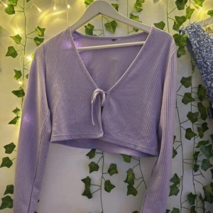 Ljuslila ribbad croppad topp - Superfin ljuslila croppad topp med långa ärmar och v-ringning. Toppen har ribbad struktur och en söt knytning framtill som ger en extra detalj. Perfekt att styla med höga jeans eller kjol.