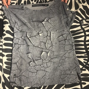 Grå topp med sprucket mönster XS - Säljer en unik grå topp från The Black Label Collection i storlek XS. Toppen har ett coolt sprucket mönster som ger en edgy vibe och är tillverkad i mjukt syntetmaterial. Rund halsringning och korta ärmar för en avslappnad look.