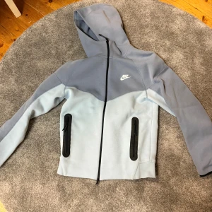 Nike Tech Fleece hoodies och byxor - Nike Tech Fleece hoodie med hel dragkedja och huva. Finns i två färger: ljusblå/grå och klarblå. Jackan har svarta detaljer vid fickorna med dragkedja, Nike-logga på bröstet och en modern, sportig look. Perfekt för chill eller träning. Fläckarna på byxorna är bara vatten. 350 kr styck för hoodiesarna och 250 för byxorna.