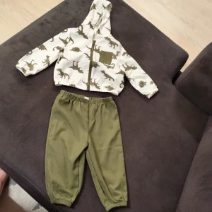 Vit vindjacka med dinosaurier från Shein - Säljer en vit vindjacka från Shein med coolt dinosauriemönster i olivgrönt. Jackan har huva, lång ärm, dragkedja framtill och en olivgrön bröstficka. Perfekt för dig som gillar lekfulla prints och vill sticka ut på förskolan.