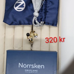 Guldring Golden Obsidian Norrsken - Snygg guldfärgad ring från Norrsken by Oriflame med en svart obsidiansten och glittrande stenar runt om. Ringen har en unik design med två dekorerade ändar, en med svart pärla och en med klar sten. Perfekt för dig som gillar statement-smycken.