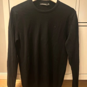 Svart stickad tröja från J.Lindeberg - Svart stickad tröja från J.Lindeberg i klassisk modell med rund hals och diskret logga på bröstet. Långärmad och mjuk, perfekt för dig som gillar stilrena och enkla plagg. Passar dig som vill ha en snygg och tidlös look. Tröjan är i storlek M.