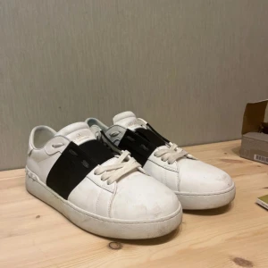 Valentino Garavani Open sneakers vit/svart - Snygga Valentino Garavani Open sneakers i vitt skinn med bred svart läderdetalj över snörningen. Klassisk rund tå, platt sula och ikoniska nitar baktill. Guldlogga på plösen. Perfekta för dig som gillar exklusiva och stilrena sneakers.