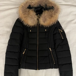 Dunjacka med äkta päls från Hollies - Snygg svart dunjacka/fjädrar från Hollies med fluffig äkta päls (tvättbjörn) runt huvan och guldfärgade dragkedjor. Jackan är figursydd, haf flera fickor med dragkedja och leopardmönstrat innerfoder. Storlek 32/XXS.