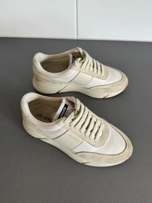 Axel Arigato - Snygga beige sneakers från Axel Arigato med chunky sula och rund tå. Skorna har en mix av mocka och textil, samt snörning framtill. Diskret logga på sidan och klassisk siluett som ger en clean vibe. Perfekta för dig som gillar minimalistisk stil.