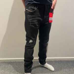 Svarta Diesel jeans med slitningar - Svarta jeans från Diesel med coola slitningar och en liten trasig detalj på låret. Klassisk femficksmodell med Diesel Industry-märke på fickan och bak. Jeansen är tillverkade i stretchigt denim för skön känsla. Jeansen är helt nya med alla lappar kvar. Säljs på grund av fel storlek. Tveka inte med att höra av er om frågor!