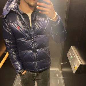 Mörkblå pufferjacka från Polo Ralph Lauren - Säljer en mörkblå pufferjacka från Polo Ralph Lauren med röd logga på bröstet. Jackan har huva, dragkedja framtill och quiltad design. Materialet är glansigt och syntetiskt, perfekt för kalla dagar. Snygg och modern look som funkar till streetwear.