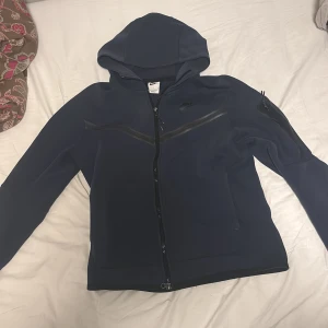 Blå Nike tech hoodie - Säljer en mörkblå Nike tech hoodie från Nike i storlek S. Jackan har huva, hel dragkedja och snygga svarta detaljer på bröstet och ärmen. Materialet är mjukt och syntetiskt, perfekt för chill eller träning. Diskret Nike-logga på bröstet och praktiska fickor framtill.