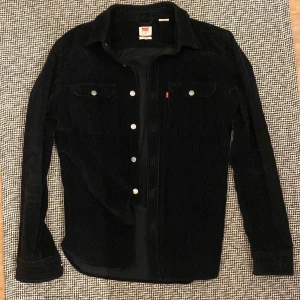 Svart manchester overshirt från Levi's - Svart overshirt från Levi's i mjuk manchester med klassisk krage och silverfärgade knappar. Två bröstfickor med knappstängning och diskret röd Levi's-tag. Perfekt att slänga över en t-shirt för en chill look.