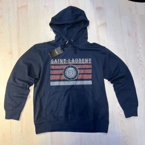 Marinblå Saint Laurent hoodie hoodie  - Snygg marinblå hoodie från Saint Laurent med tryck i rött och vitt framtill. Tröjan har huva med snörning, stor magficka och långa ärmar. Materialet är mjuk bomull och passformen är normal. Perfekt för dig som gillar streetwear och vill sticka ut med ett coolt märke. 0bs lnte 0rglnal 