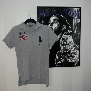 Grå piké från Polo Ralph Lauren - Säljer en grå pikétröja från Polo Ralph Lauren i storlek S. Tröjan har klassisk krage, korta ärmar och broderad blå polospelare på bröstet. USA-flagga och texten 'USA R. Lauren' på vänster sida. Custom fit-modell i mjuk bomull.