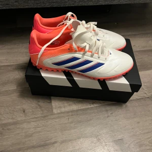 Adidas Copa Pure fotbollsskor vit/orange - Säljer ett par Adidas Copa Pure fotbollsskor i vitt med blå ränder och orange detaljer. Skorna har snörning, platt sula med dobbar och är tillverkade i syntetmaterial. Perfekta för dig som vill sticka ut på planen med stilrena och färgstarka skor.