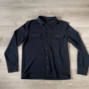 Mörkblå overshirt från Carry Our Passion - Mörkblå overshirt från Carry Our Passion i storlek M. Skjortan har klassisk krage, två stora bröstfickor med lock och knappar hela vägen framtill. Materialet känns mjukt och har en stilren look som passar till många outfits. Skriv för fler bilder med mera