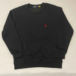 Svart sweatshirt från Polo Ralph Lauren - Svart sweatshirt från Polo Ralph Lauren med klassisk rund hals och lång ärm. Tröjan har en röd broderad logga på bröstet och ribbade muddar vid ärmslut och nederkant. Perfekt för dig som gillar stilren och enkel design.