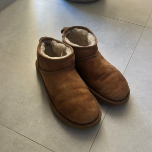 Bruna miniuggs - Mysiga bruna UGG boots med rund tå och platt sula. Utsidan är i mjuk mocka och insidan är fodrad med fluffigt som håller fötterna varma. Klassisk UGG-logga baktill och praktisk flik för att enkelt ta på skorna. Perfekta för kyliga dagar. Fint skick men väl använda.