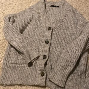 Grå stickad kofta med knappar - Mysig grå stickad kofta från FIVE UNITS i storlek XS. Koftan har grov ribbstickning, stora svarta knappar framtill och två praktiska fickor. Perfekt för lager-på-lager och chill stil. Långärmad och med breda muddar vid ärmslut och nederkant.