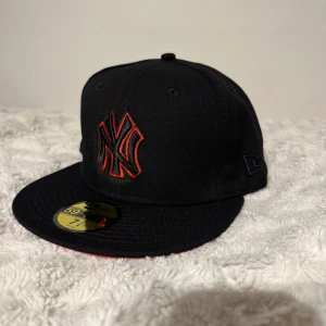 Svart New Era Yankees 59FIFTY keps - Snygg svart New Era 59FIFTY keps med New York Yankees-logga i svart och röd brodyr framtill. På sidan finns ett broderat 1999 World Series-märke i rött och svart. Klassisk rak skärm och MLB-logga baktill. Perfekt för dig som gillar streetwear och sport.