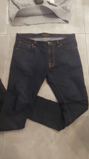 Mörkblå Nudie Jeans 32/34 - Säljer ett par mörkblå jeans från Nudie Jeans i klassisk rak modell. Jeansen har orange kontrastsömmar, fem fickor och Nudie-logga på bakfickan. Tillverkade i slitstarkt bomullsjeansmaterial. Perfekt för dig som gillar stilrena och tidlösa jeans.