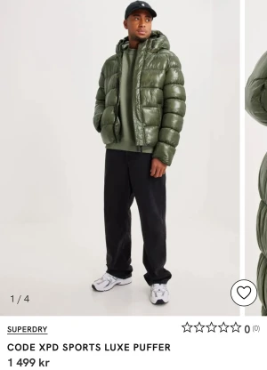 Grön pufferjacka från Superdry - Säljer en grön pufferjacka från Superdry med huva och glansig finish. Jackan har breda, quiltade sektioner och dragkedja framtill. Perfekt för dig som gillar streetwear och vill ha en varm jacka med cool look. Passar till både hoodie och t-shirt.