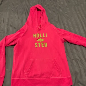 Rosa hoodie från Hollister med huva - Säljer en snygg rosa hoodie från Hollister med limegröna broderade bokstäver på bröstet. Tröjan har huva med snörning och en stor magficka. Perfekt för dig som gillar streetstyle och vill ha något bekvämt och färgstarkt.