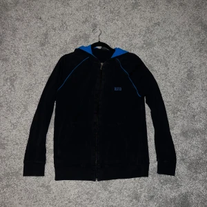 Svart hoodie från BOSS med blå detaljer - Svart hoodie från BOSS med blå huva och blå detaljer längs axlarna. Tröjan har dragkedja framtill, fickor och BOSS-logga på bröstet. Perfekt för en avslappnad och sportig stil.