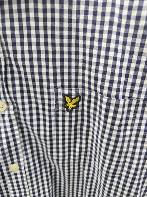 Rutig blåvit skjorta Lyle & Scott - Slim fit skjorta från Lyle & Scott i blåvitt rutigt mönster. Klassisk krage med knappar hela vägen och broderad gul logga på bröstet. Långärmad och tillverkad i mjuk bomull, perfekt för dig som gillar stilren och enkel design.