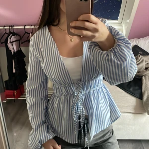 Blårandig blus med snörning - Säljer en blårandig blus med v-ringning och snörning i midjan. Blusen har långa ärmar och ett luftigt, loose fit. Perfekt för dig som gillar en chill och stilren look med lite extra detaljer.
