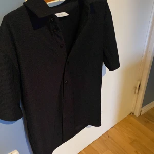 Svart ribbad skjorta från Grunt - Svart skjorta från Grunt i storlek L med ribbad struktur och klassisk krage. Skjortan har korta ärmar och knappar framtill. Materialet känns mjukt och har en snygg textur som ger extra vibe till outfiten. Passformen är relaxed/baggy och har använts 1 gång till en skolavslutning. Man kan matcha den med svarta linnebyxor och en vit tshirt under för lite finare outfit på semester, sommaren eller bara lite finare stunder.