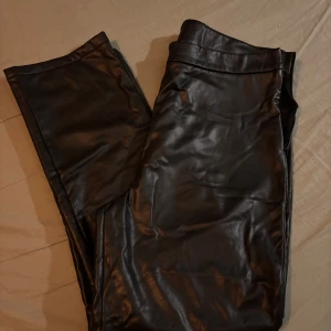 Svarta skinnbyxor med rak passform - Snygga svarta skinnbyxor med rak passform och klassisk midja. Byxorna har en stilren look med slät yta och är tillverkade i syntetmaterial som ger en cool läderkänsla. Perfekta för dig som vill ha en edgy vibe i garderoben.