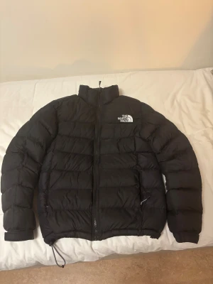 Svart pufferjacka från The North Face - Svart pufferjacka från The North Face med hög krage och synligt logotryck på bröstet. Jackan har dragkedja framtill, snörning i nederkant och två sidofickor. Den är quiltad och har långa ärmar, perfekt för kalla dagar.