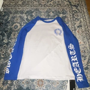 Chrome Hearts blåvit långärmad tröja - Snygg långärmad tröja från Chrome Hearts med blå ärmar och vit kropp. Tryck med Chrome Hearts-logga på bröstet och stora bokstäver på ryggen. Ärmarna har coolt tryck längs hela längden. Perfekt för dig som gillar streetwear och vill sticka ut.
