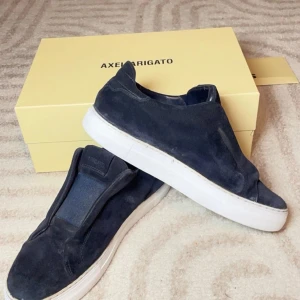 Mörkblå mocka sneakers Axel Arigato - Snygga mörkblå sneakers från Axel Arigato i mjuk mocka med vit platt sula. Skorna har en minimalistisk design utan snörning och elastiska paneler för enkel påtagning. Perfekta för dig som gillar stilrena och trendiga skor med premiumkänsla.