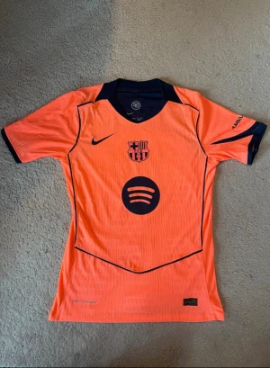 Fc Barcelona Pedri Nike orange matchtröja Size M - Säljer en FC Barcelona matchtröja med Pedri 8 på ryggen. Tröjan är orange med mörkblå detaljer, klubbmärke och sponsortryck från Spotify och UNHCR. Tillverkad i lätt funktionsmaterial, perfekt för fotboll eller att bära som supporter.