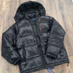 Svart pufferjacka från Polo Ralph Lauren - Svart pufferjacka från Polo Ralph Lauren med huva och dragkedja. Jackan har en glansig finish, två sidofickor med dragkedja och den klassiska röda Polo-loggan broderad på bröstet. Perfekt för kalla dagar med sin tjocka vaddering och justerbar nederkant.
