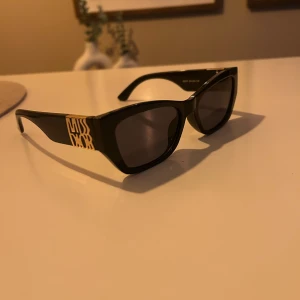 Svarta Miss Dior solglasögon - Snygga svarta solglasögon från Miss Dior med rektangulär form och breda skalmar. På sidan finns en cool guldfärgad Miss Dior-logga som ger extra lyxig vibe. Glasen är mörka och bågarna är tillverkade i blank syntet. Perfekt accessoar för att höja din stil.