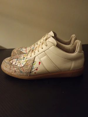 Maison margiela  - Säljer ett par vita sneakers med beige mockadetaljer och coola färgstänk i rött, blått, gult och grönt. Skorna har snörning, platt sula och rund tå. Ovandelen är i skinn och mocka, vilket ger en schysst kontrast. Perfekt för dig som vill sticka ut med din stil. 