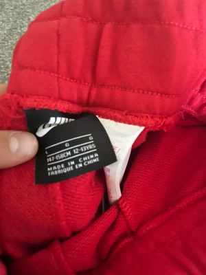 Nike röda mjukisbyxor med färgblock - Snygga röda mjukisbyxor från Nike med coola färgblock i svart och vitt på benen. Byxorna har elastisk midja med snörning och är gjorda i mjukt bomullsmaterial. Perfekta för chill eller träning, med en loose passform och sportig vibe.