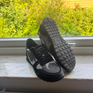 Valentino Garavani Rockrunner sneakers - Svarta och grå Valentino Garavani Rockrunner sneakers med coolt kamouflagemönster. Skorna har snörning, platt sula och rund tå. Materialmix med mesh och mockadetaljer ger en schysst streetkänsla. Kommer med originalkartong och röd dustbag.