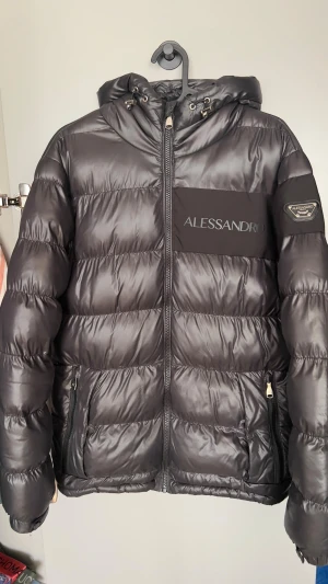 Svart pufferjacka Alessandro Zavetti - Svart pufferjacka från Alessandro Zavetti med snyggt glansigt tyg och logga på bröstet och ärmen. Jackan har huva med snörning, två fickor med dragkedja och markerade sömmar.