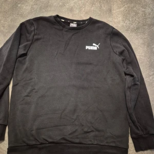 Svart sweatshirt från Puma - Svart sweatshirt från Puma med klassisk rund hals och vit logotyp på bröstet. Tröjan har långa ärmar och ribbade muddar vid ärmslut och nederkant. 