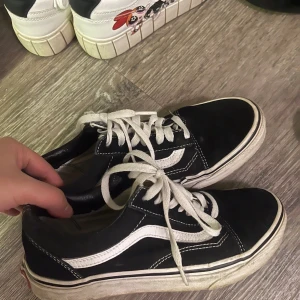 Svarta Vans Old Skool sneakers - Klassiska Vans Old Skool sneakers i svart med vit sula och vit sidostripes. Skorna har snörning och rund tå, tillverkade i skinn och textil. Perfekt för dig som gillar streetstyle och vill ha en tidlös look.