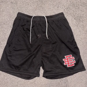 Svarta shorts från Eric emanuel - Svarta mesh shorts från EE, med vit snörning i midjan och ett stort rött och vitt EE-logo tryck på vänster ben. Luftigt material som passar perfekt för sport eller chill. Elastisk midja för skön passform. Tillverkade i New York. Storlek L