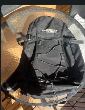 Svart ryggsäck från Everest - Snygg svart ryggsäck från Everest med praktiska spännen och snörning framtill. Perfekt för skolan eller utflykter, med flera fack och justerbara axelremmar. Enkel och stilren design som passar till det mesta.