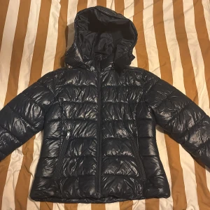 Mörkblå pufferjacka från 157 - Säljer en mörkblå pufferjacka från 157 med huva och två snedställda dragkedjefickor. Jackan har quiltad design och är tillverkad i syntetmaterial. Perfekt för kalla dagar och har en clean look med hel dragkedja framtill.