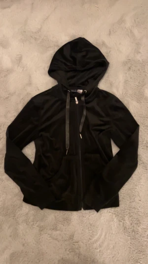 Svart hoodie i sammet med huva - Svart hoodie i mjuk sammet med huva och snörning. Tröjan har dragkedja framtill och långa ärmar, samt en stor ficka på magen. Perfekt för chill dagar och när du vill vara extra comfy.