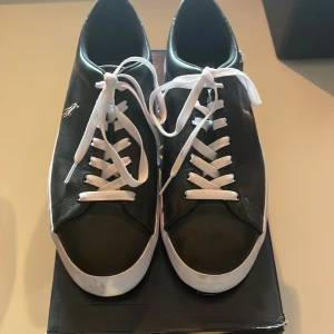Svarta sneakers från Polo Ralph Lauren - Snygga svarta sneakers från Polo Ralph Lauren med vit sula och vita snören. Skorna har en klassisk låg profil och är tillverkade i skinn med Polo-loggan broderad på sidan. Yttersulan är brun med tydligt mönster för bra grepp. Perfekt för dig som gillar stilrena och tidlösa sneakers. Skorna är i princip ny skick. Storlek 44 men passar även 43 och 45. Nypris 1400 mitt pris 700